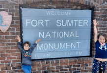 Two children excited with their hands up, standing on either side of the Fort Sumter National Monument sign.