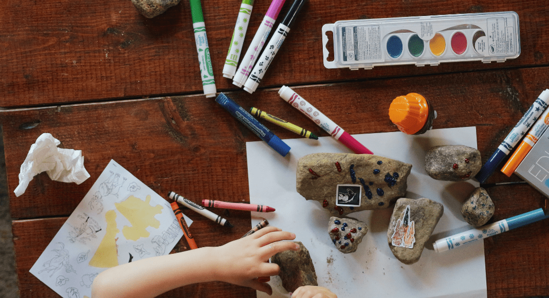 A table covered with rocks and art supplies, with a child drawing on a piece of paper.