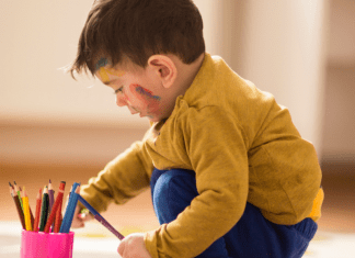 World Poetry Day: a preschool-aged boy crouches down on a giant piece of paper laying on the floor. He has a cup full of colored pencils with one in hand, and splashes of color scribbled on his cheek.